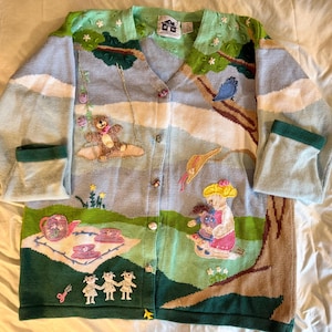 May include: A whimsical, long-sleeve cardigan sweater with a V-neck. The sweater features a colorful, embroidered scene with teddy bears, a tea party, and a bluebird. The color palette includes shades of blue, green, pink, and brown.