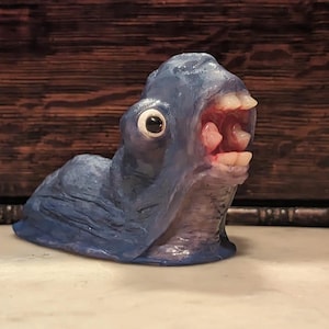 Puede incluir: Una escultura de cerámica azul pintada a mano de una criatura con una boca grande y dientes. La criatura tiene un solo ojo grande y una superficie texturizada.