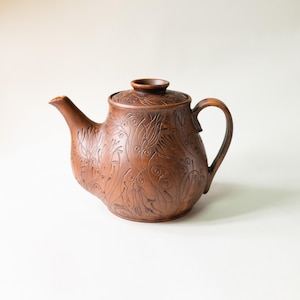 Puede incluir: Una tetera de arcilla marrón con un cuerpo redondeado y una tapa a juego. La superficie está decorada con intrincados patrones florales y de remolinos oscuros grabados que cubren todo el exterior, incluyendo el pico y el asa curva.