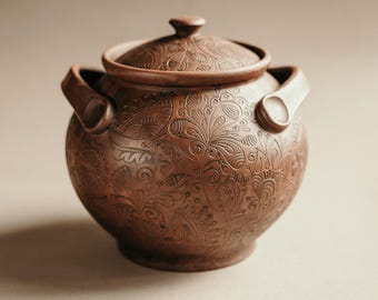 May include: A brown ceramic pot with a lid and two handles. The pot features an intricate floral pattern carved into its surface. The lid has a small knob for easy removal. The pot is likely used for cooking or storage.