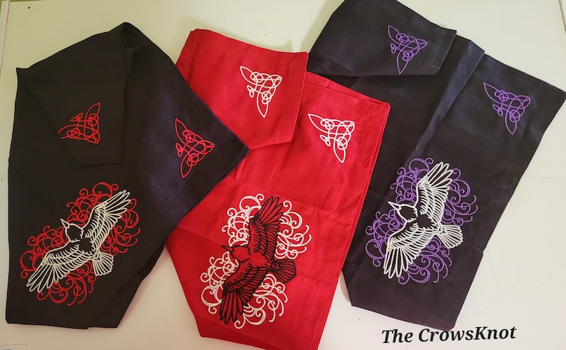 K&ouml;nnte beinhalten: Drei Stoffquadrate mit gestickten Designs. Das Quadrat links ist schwarz mit einem wei&szlig;en Raben und roten Schn&ouml;rkeln. Das Quadrat in der Mitte ist rot mit einem schwarzen Raben und wei&szlig;en Schn&ouml;rkeln. Das Quadrat rechts ist schwarz mit einem wei&szlig;en Raben und lila Schn&ouml;rkeln. Jedes Quadrat hat ein kleines keltisches Knotendesign in der Ecke.
