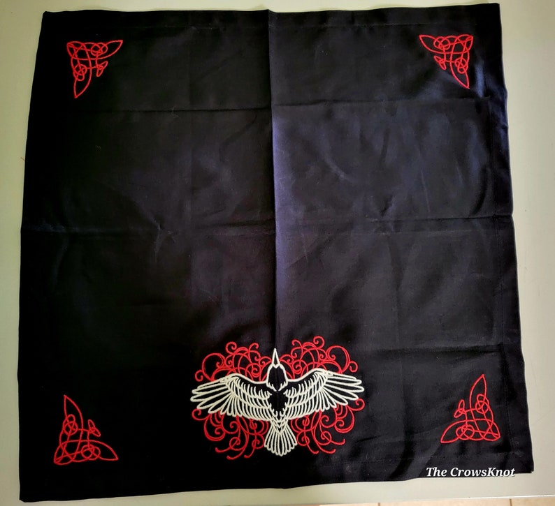 K&ouml;nnte beinhalten: Schwarzer Stofftischdecke mit roten und wei&szlig;en gestickten keltischen Knotenmustern und einem wei&szlig;en Raben in einem roten Blumendesign. Der Text "The CrowsKnot" befindet sich in der rechten unteren Ecke.