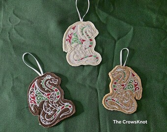 May include: Three dragon-shaped ornaments with white ribbon hangers. The ornaments are embroidered with a dragon design in shades of brown, beige, red, and green. The background is a dark green fabric. The text "The CrowsKnot" is visible.