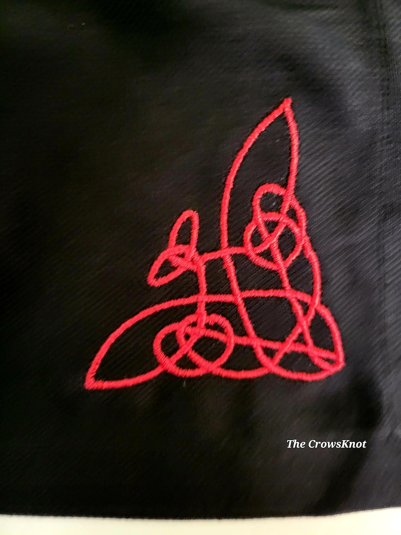 K&ouml;nnte beinhalten: Ein rotes, gesticktes keltisches Knotendesign auf einem schwarzen Stoffhintergrund. Der Text "The CrowsKnot" ist am unteren Rand des Bildes sichtbar.