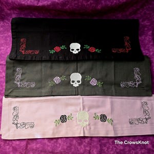 May include: Three rectangular decorative cloths in black, gray-green, and pink, stacked on top of each other. Each cloth features an embroidered skull, roses, and corner flourishes. The bottom right corner of the image has the text "The CrowsKnot".