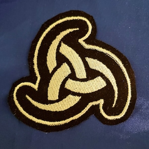 May include: A black and gold embroidered patch with a stylized knot design. The patch features a three-looped knot with a white outline. The text "The CrowsKnot" is visible below the patch.