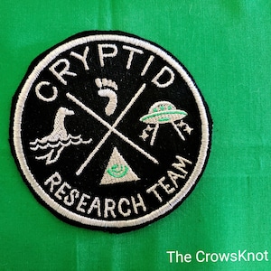 May include: Black and white embroidered patch with the text "Cryptid Research Team" in a circle. The patch features a footprint, a UFO, a creature from the water, and a triangle with an eye inside.