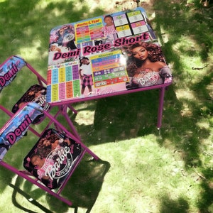 Puede incluir: Un conjunto de mesa y dos sillas rosas para niños con temática Barbie, sobre césped. La mesa presenta el texto "Demi Rose Short" y gráficos educativos. Las sillas tienen "Demi" en los respaldos y gráficos de Barbie en los asientos.