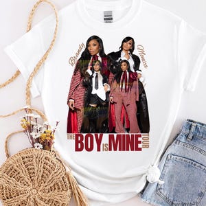 Pode incluir: Camiseta branca com um gráfico de Brandy e Monica, e o texto "The Boy Is Mine Tour" em vermelho. A imagem mostra as cantoras em várias roupas, incluindo ternos e casacos.