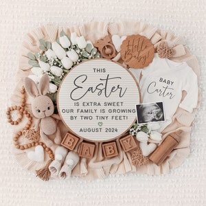 May include: A wooden sign with the text "This Easter is extra sweet our family is growing by two tiny feet! August 2024" surrounded by a white baby onesie, a wooden rattle, a crocheted bunny, wooden blocks spelling "baby", and a white flower arrangement.