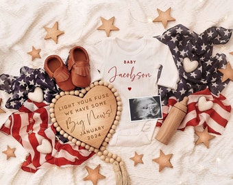 May include: A wooden heart-shaped sign with the text "Light your fuse we have some Big News! January 2024" surrounded by red, white, and blue fabric, baby booties, and a white onesie with the text "Baby Jacobson" in red.