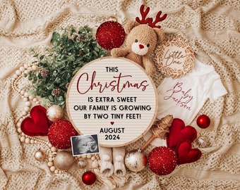 May include: A wooden sign with the text "This Christmas is extra sweet our family is growing by two tiny feet! August 2024" surrounded by red and gold baubles, a stuffed reindeer, a baby onesie, and a small Christmas tree.