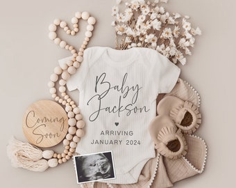 Puede incluir: Un body blanco con el texto "Baby Jackson Arriving January 2024" en él. El body está rodeado de una guirnalda de cuentas de madera, un letrero de madera que dice "Coming Soon", flores secas, una manta para bebé y un par de zapatos para bebé.