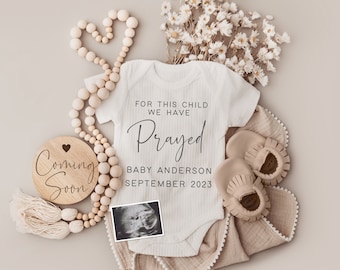 Peut inclure: Une grenouillère blanche avec le texte "For this child we have prayed" et "Baby Anderson September 2023". La grenouillère est entourée de perles en bois, de fleurs séchées, d'une pancarte en bois qui dit "Coming Soon" et d'une paire de chaussons beige pour bébé.