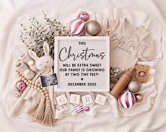 May include: A white letter board with the text "This Christmas will be extra sweet our family is growing by two tiny feet! December 2023".  The board is surrounded by pink and gold ornaments, a baby onesie, a wooden rattle, and a pair of baby booties.