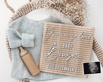 Puede incluir: Un tablero de letras de madera con el texto "You are so loved Baby Benson Due December 22" en letras blancas. Un pequeño suéter de punto y un biberón de madera están en una cesta de mimbre junto al tablero de letras. Una imagen de ultrasonido está en la esquina inferior derecha de la imagen.