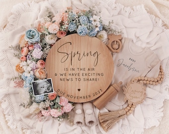 May include: A wooden announcement sign with the text "Spring is in the air & we have exciting news to share! Due November 2023" is surrounded by flowers, a baby onesie, and a sonogram. A wooden baby bottle and a "Coming Soon" sign are also present.