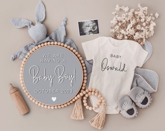 May include: A gray wooden circle sign with white text that reads "We are having a Baby Boy! October 2023". A white onesie with the text "Baby Oswald" is next to the sign. There is a baby rattle, a pair of baby shoes, and a baby blanket in the image.