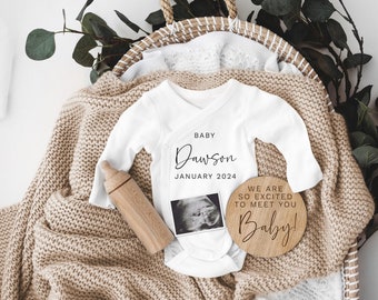 Könnte beinhalten: Ein weißer Baby-Strampler mit dem Text "Baby Dawson Januar 2024" und einem kleinen Ultraschallbild. Eine Holz-Babyflasche und ein Holzschild mit dem Text "We are so excited to meet you Baby!" sind ebenfalls auf dem Bild zu sehen.