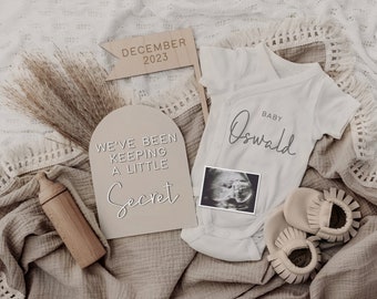 Könnte beinhalten: Eine geschlechtsneutrale Baby-Ankündigung mit einem weißen Strampler, auf dem "Baby Oswald" steht, und einem Schild mit der Aufschrift "We've been keeping a little secret". Außerdem sind ein Holz-Babyfläschchen, ein Ultraschallbild und ein Paar Babyschuhe zu sehen.