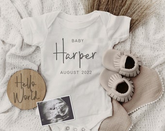 Könnte beinhalten: Ein weißer Baby-Strampler mit dem Text "BABY Harper AUGUST 2022" darauf. Neben dem Strampler befindet sich ein Holzschild mit dem Text "Hello World". Auf einer weißen Strickdecke liegen ein kleines Ultraschallbild und ein Paar beige Baby-Mokassins.
