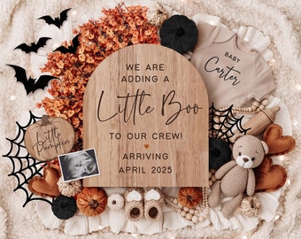 May include: A wooden sign with the text "We are adding a little boo to our crew! Arriving April 2025" and a baby onesie with the text "Baby Carter". The sign is surrounded by Halloween decorations, including a spiderweb, bats, pumpkins, and a teddy bear.