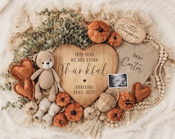 Könnte beinhalten: Ein herzförmiges Schild aus Holz mit dem Text "This year we are extra thankful" und "Arriving April 2025". Neben dem Herzen befindet sich ein kleineres Holzschild mit dem Text "Baby Carter". Die Schilder sind von Herbstdekorationen umgeben, darunter ein kleiner Stoffteddybär, ein Holzschild in Form eines Kürbisses und eine Schnur aus Holzperlen.
