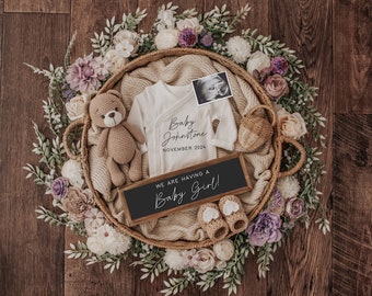 May include: A wicker basket filled with a white onesie, a teddy bear, a small wooden sign that reads "We are having a Baby Girl!" and a black and white ultrasound image. The basket is surrounded by a wreath of white and purple flowers.