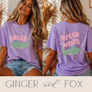 May include: A lilac t-shirt featuring a cartoon alligator design. The front of the shirt says "later" above the alligator, and the back says "after while". The alligator is green with pink accents and is surrounded by yellow stars.