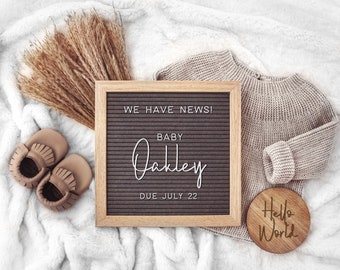 Könnte beinhalten: Baby-Ankündigung in neutralen Tönen. Mit einer Buchstaben-Tafel mit dem Text "WE HAVE NEWS! BABY Oakley DUE JULY 22". Die Szene umfasst einen Strickpullover, Babyschuhe, eine Holzscheibe mit "Hello World" und getrockneten Weizen.