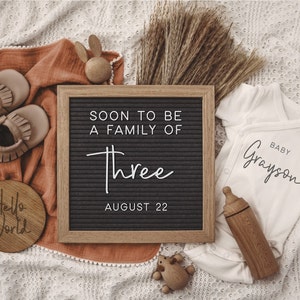 Może przedstawiać: Czarna tablica z literami z tekstem "Soon to be a family of three August 22". Obok tablicy z literami znajduje się biały śpioszek z tekstem "Baby Grayson". Są też buciki dla niemowląt, drewniana zabawka i drewniany znak z napisem "Hello World".