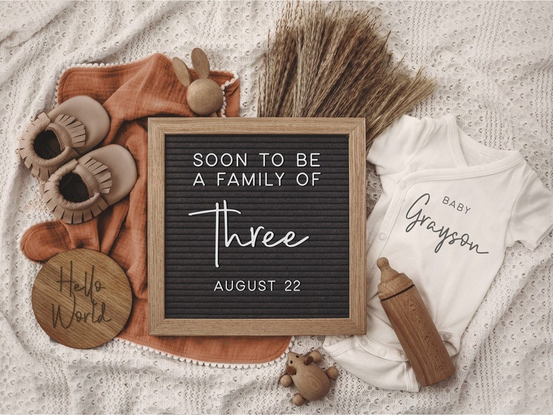 Może przedstawiać: Czarna tablica z literami z tekstem "Soon to be a family of three August 22". Obok tablicy z literami znajduje się biały śpioszek z tekstem "Baby Grayson". Są też buciki dla niemowląt, drewniana zabawka i drewniany znak z napisem "Hello World".
