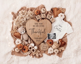 Könnte beinhalten: Ein herzförmiges Schild aus Holz mit dem Text "This year we are extra thankful February 2025" umgeben von Herbstdekorationen, darunter ein Baby-Strampler mit dem Text "Baby Carter", ein kleiner Teddybär und ein Kürbis mit dem Text "Coming Soon".