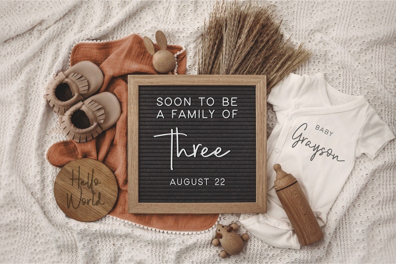 Może przedstawiać: Czarna tablica z białymi literami, kt&oacute;re tworzą napis "Soon to be a family of three August 22". Obok tablicy znajduje się biały śpioszek z napisem "Baby Grayson". Na zdjęciu widać brązowe buciki dla dziecka, drewnianą zabawkę i drewniany znak z napisem "Hello World".