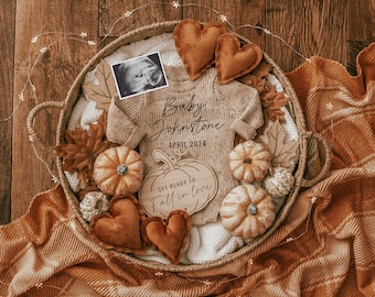 May include: A wicker basket filled with fall-themed decor, including pumpkins, leaves, and a baby onesie. The onesie has the text "Baby Johnstone April 2024" printed on it. A wooden sign reads "Get ready to fall in love".