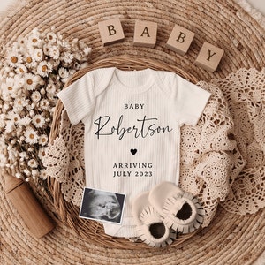 K&ouml;nnte beinhalten: Ein wei&szlig;er Strampler mit dem Text "BABY Robertson ARRIVING JULY 2023" und einem kleinen Herz. Der Strampler ist umgeben von Holzbl&ouml;cken, die "BABY" buchstabieren, einer H&auml;keldecke und einem Strau&szlig; getrockneter Blumen.