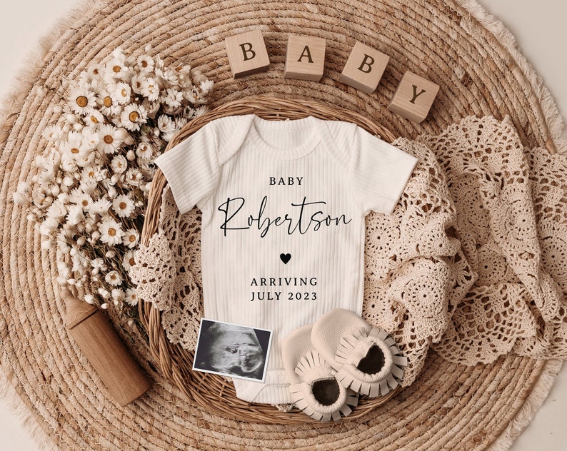 K&ouml;nnte beinhalten: Ein wei&szlig;er Strampler mit dem Text "BABY Robertson ARRIVING JULY 2023" und einem kleinen Herz. Der Strampler ist umgeben von Holzbl&ouml;cken, die "BABY" buchstabieren, einer H&auml;keldecke und einem Strau&szlig; getrockneter Blumen.