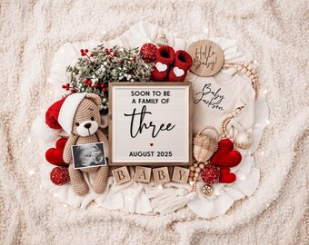 May include: A Christmas-themed pregnancy announcement with a letter board that reads "Soon to be a family of three August 2023". There is a teddy bear wearing a Santa hat, a baby onesie, and a wooden sign that says "Hello Baby!"