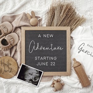 May include: A black letter board with the text "A new adventure starting June 22". A wooden baby rattle, a pair of brown baby moccasins, a wooden baby bottle, a white onesie with the text "Baby Garcia", a wooden sign with the text "Hello World", and a black and white ultrasound image are arranged on a white crocheted blanket.