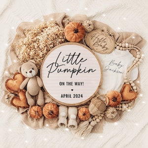 May include: A white wooden sign with the text "Little Pumpkin On The Way! April 2024" surrounded by fall-themed decorations, including a teddy bear, pumpkins, and a baby onesie with the text "Baby Jackson".