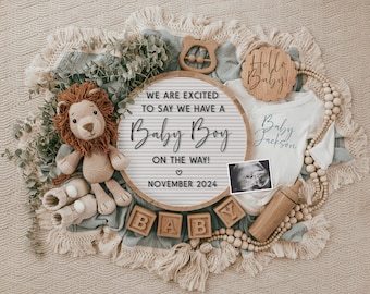 Op de afbeelding: Een houten bord met de tekst "WE ARE EXCITED TO SAY WE HAVE A Baby Boy ON THE WAY! ♡ NOVEMBER 2024" omringd door babyartikelen zoals een knuffel leeuw, een rompertje, houten blokken en een houten rammelaar.