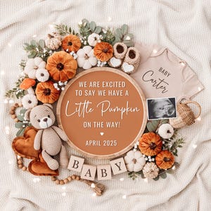 Op de afbeelding: Een houten bord met de tekst "We are excited to say we have a little pumpkin on the way! April 2025" omringd door herfstdecoraties, waaronder pompoenen, bladeren en een teddybeer. Het bord ligt op een wit deken.