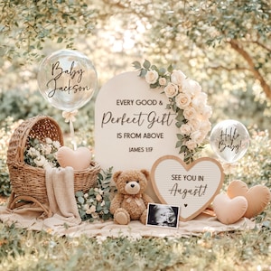 Puede incluir: Una escena de anuncio de bebé con una cuna de mimbre llena de flores, un oso de peluche y decoraciones en forma de corazón. Un letrero dice "Todo buen y perfecto regalo viene de arriba". Globos dicen "Baby Jackson" y "Hello Baby!"