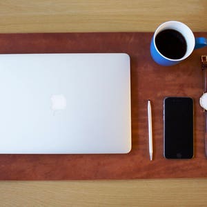 Puede incluir: Una alfombrilla de escritorio de cuero marr&oacute;n con un port&aacute;til plateado, una taza de caf&eacute; azul, un bol&iacute;grafo plateado, un tel&eacute;fono inteligente negro y un reloj de pulsera de cuero marr&oacute;n con una esfera blanca.