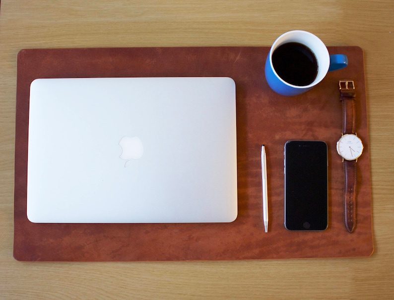 Puede incluir: Una alfombrilla de escritorio de cuero marr&oacute;n con un port&aacute;til plateado, una taza de caf&eacute; azul, un bol&iacute;grafo plateado, un tel&eacute;fono inteligente negro y un reloj de pulsera de cuero marr&oacute;n con una esfera blanca.