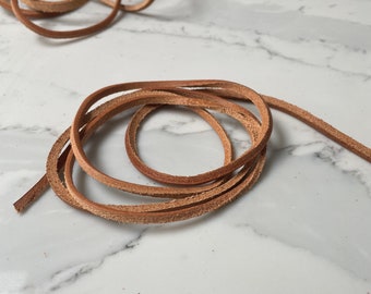 May include: Close-up of several strands of brown leather cord, coiled and scattered. The leather appears to be natural in color, with visible texture and slight variations in tone. The cords are arranged on a white and gray marble surface.
