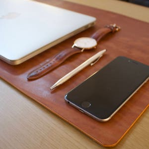 Puede incluir: Una alfombrilla de escritorio de cuero marr&oacute;n con un port&aacute;til, un reloj, un bol&iacute;grafo y un tel&eacute;fono inteligente encima.