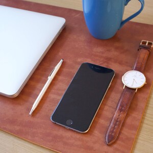 Puede incluir: Una alfombrilla de escritorio de cuero marr&oacute;n con un port&aacute;til, un tel&eacute;fono inteligente, un bol&iacute;grafo y un reloj de pulsera con esfera blanca y correa de cuero marr&oacute;n.