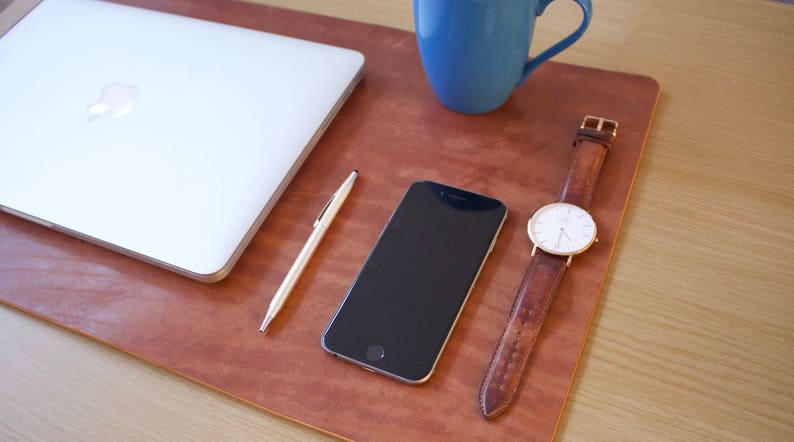 Puede incluir: Una alfombrilla de escritorio de cuero marr&oacute;n con un port&aacute;til, un tel&eacute;fono inteligente, un bol&iacute;grafo y un reloj de pulsera con esfera blanca y correa de cuero marr&oacute;n.
