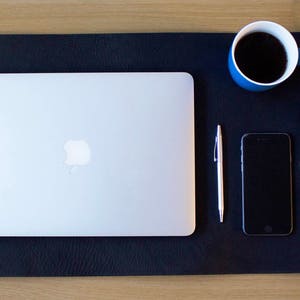 Puede incluir: Una alfombrilla de escritorio de cuero negro con un port&aacute;til, un tel&eacute;fono inteligente, un bol&iacute;grafo, una taza de caf&eacute; azul y un reloj de pulsera con una correa de cuero marr&oacute;n.